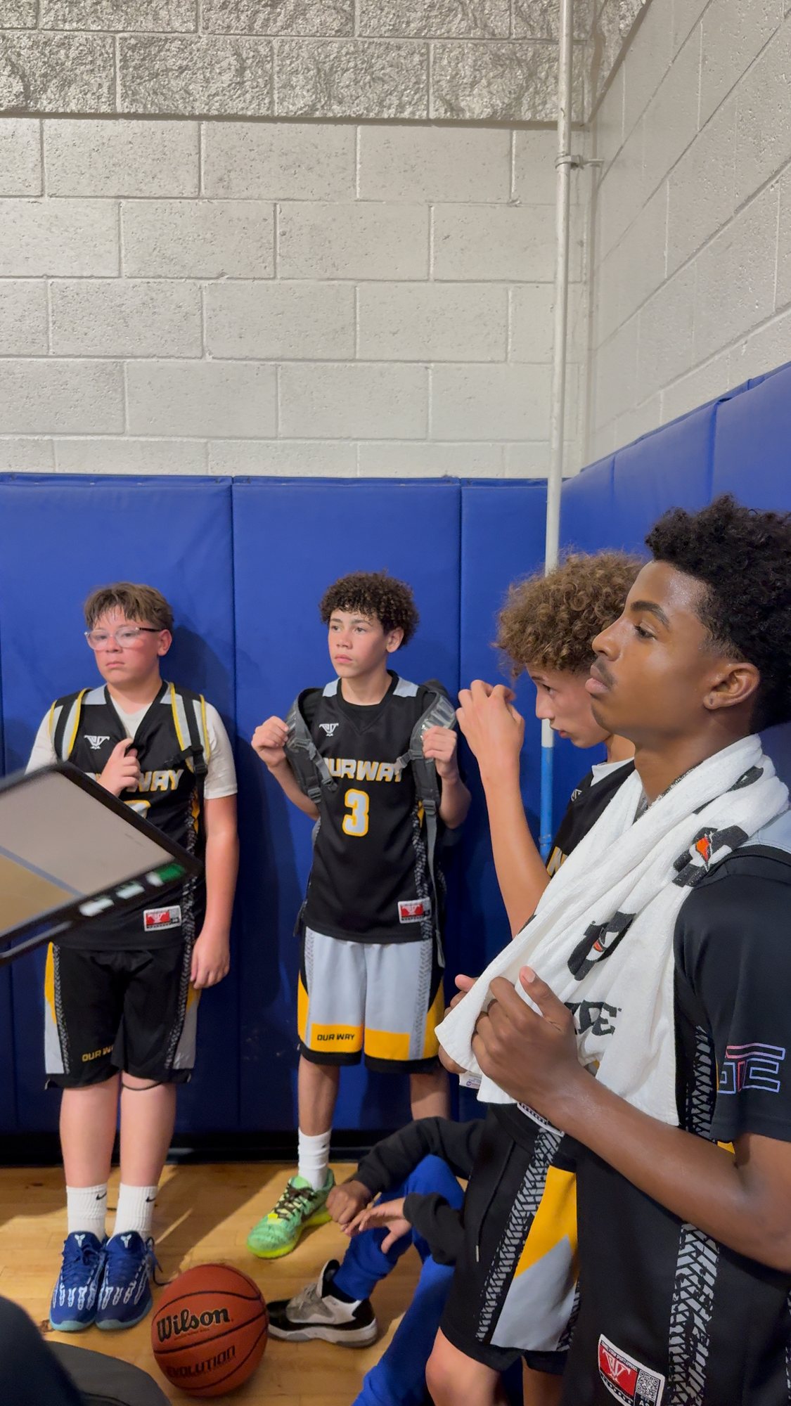OW players standing along the padded gym wall beside a coach’s clipboard